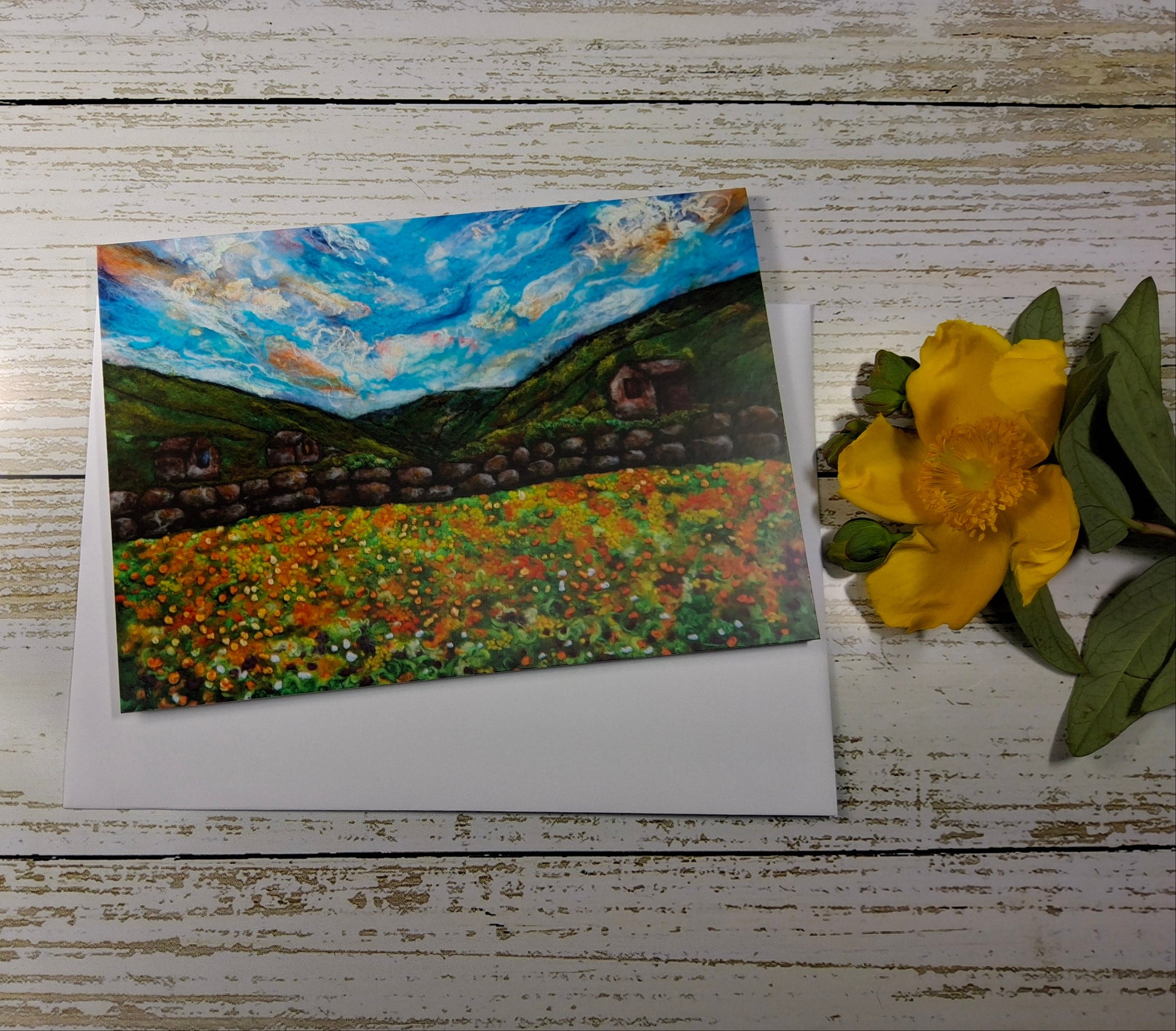 A square art print or card featuring an impressionistic landscape painting with a blue sky, green hills, and a colorful flower field, displayed on a white surface next to a yellow flower on a whitewashed wood background.