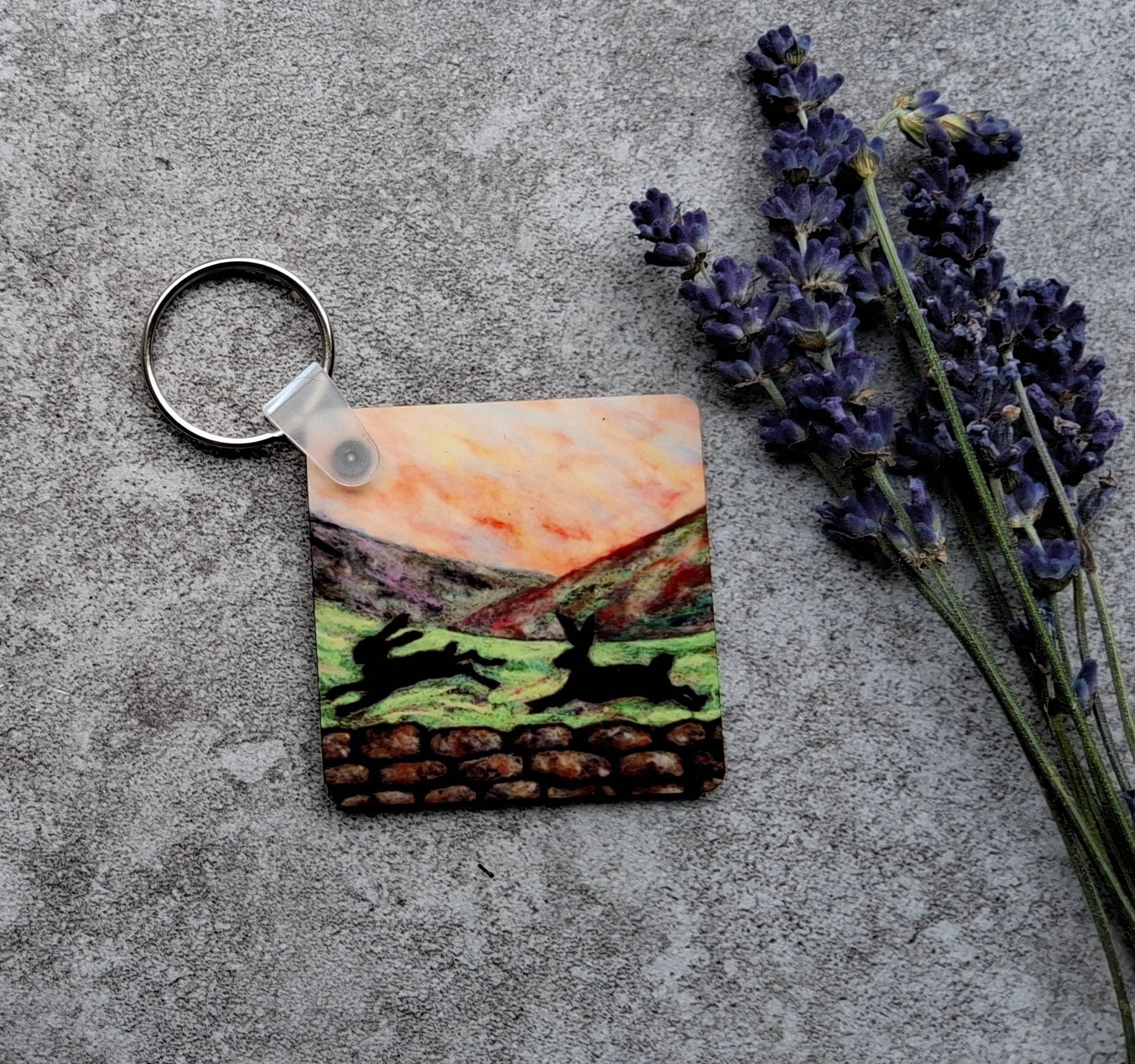 A square acrylic keychain featuring a colorful landscape illustration of deer in a field, resting on a textured gray concrete surface next to dried lavender sprigs.