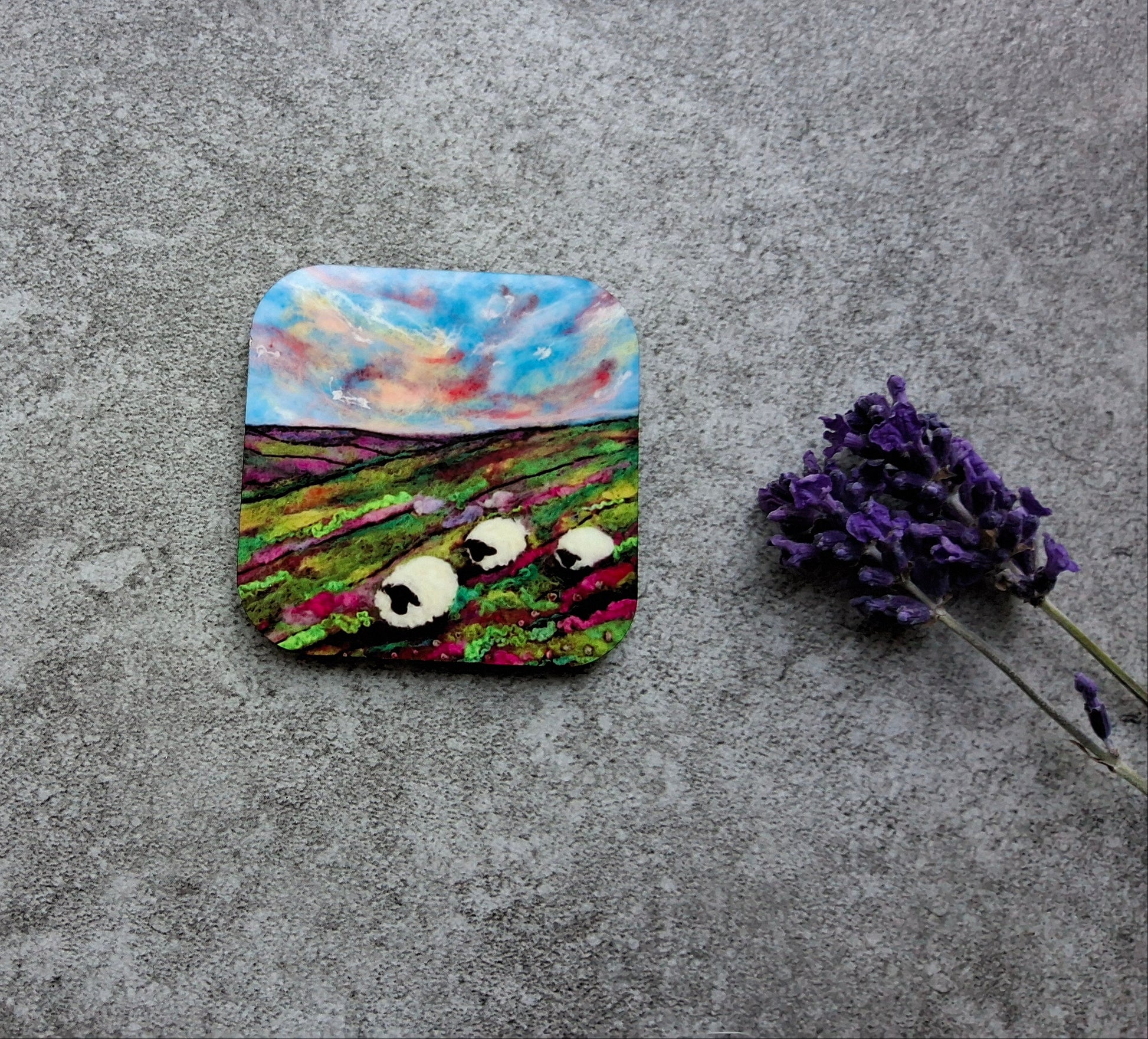 A square coaster with rounded corners featuring a painted landscape of a lavender field with sheep under a cloudy sky, displayed on a textured gray surface next to a sprig of dried lavender.