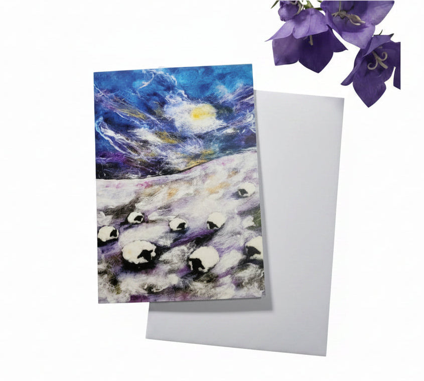 A square greeting card featuring a colorful painting of penguins in a snowy landscape under a starry night sky, placed on a white envelope over a light wood background with purple flowers.