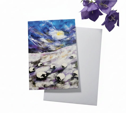A square greeting card featuring a colorful painting of penguins in a snowy landscape under a starry night sky, placed on a white envelope over a light wood background with purple flowers.