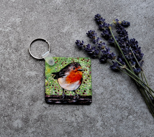 A square keychain featuring a painted robin bird on a green textured background, attached to a silver split ring and placed on a gray concrete surface next to dried lavender sprigs.