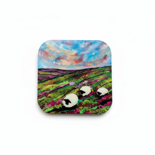 A square coaster with rounded corners featuring a colorful painted landscape of sheep in a field under a blue sky, placed on a textured gray surface next to dried purple lavender.