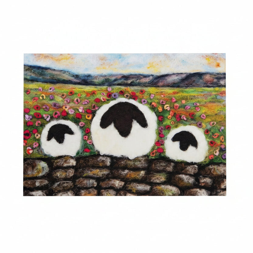 A square art print or card featuring a painted landscape with three white sheep in front of a stone wall, placed on a white wooden surface next to a small purple flower sprig.