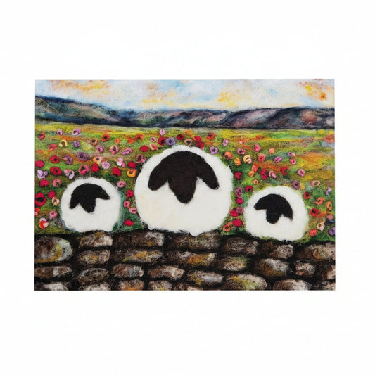 A square art print or card featuring a painted landscape with three white sheep in front of a stone wall, placed on a white wooden surface next to a small purple flower sprig.