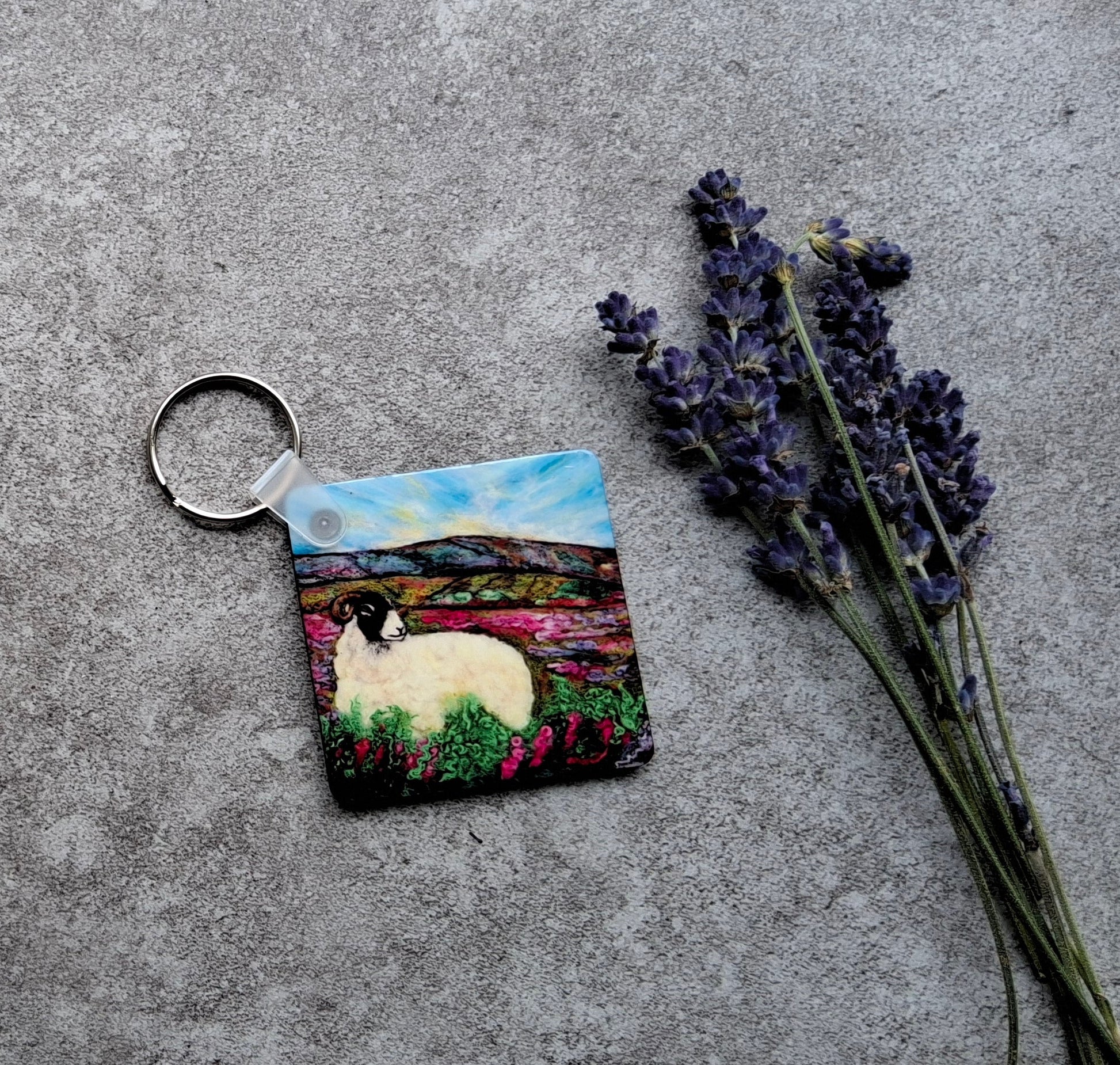 A square painted keychain featuring a white sheep in a colorful landscape, attached to a silver metal ring, styled on a textured gray concrete surface with dried lavender and yellow flowers.