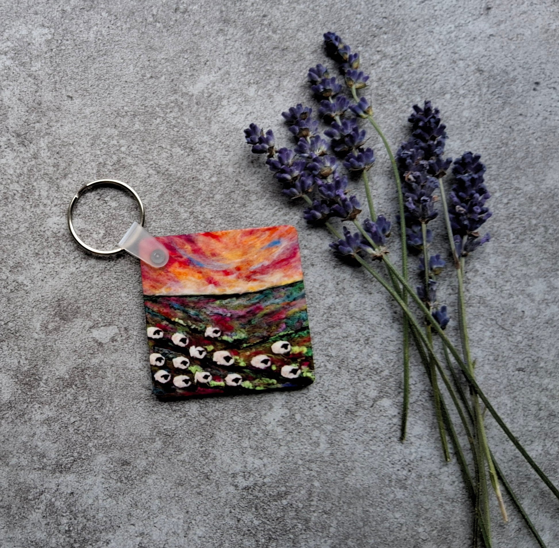A square acrylic keychain featuring a colorful landscape painting of a field under a sunset sky, shown on a textured gray concrete surface next to sprigs of lavender.