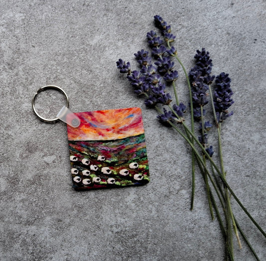 A square acrylic keychain featuring a colorful landscape painting of a field under a sunset sky, shown on a textured gray concrete surface next to sprigs of lavender.
