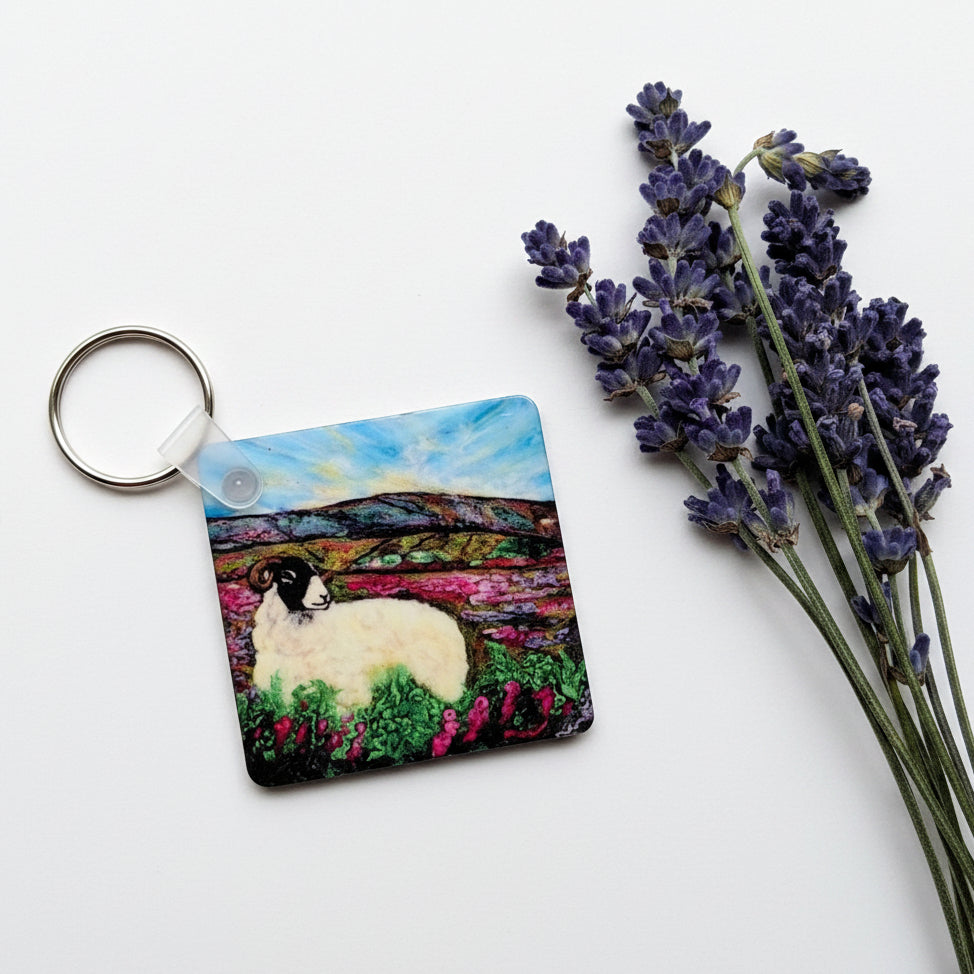 A square keychain featuring a colorful painted scene of a white sheep in a field under a blue sky, placed on a textured gray surface next to dried lavender sprigs.