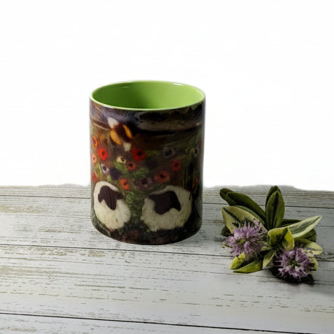 A cylindrical ceramic mug with a colorful owl face design and a bright green interior, placed on a white wood-grain tabletop next to a small sprig of flowers, with a desk lamp and a large marble-patterned vase in the background.