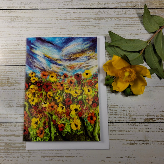 A vertical art print or card featuring an impressionistic painting of a flower field under a swirling blue sky, styled on a white wood background next to a yellow flower.