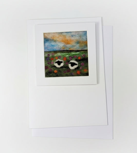 A small framed art print featuring two white birds with red accents against a blue and orange sky, displayed on a white card and envelope over a rustic whitewashed wooden surface with decorative greenery.