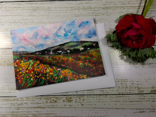 A square landscape painting of a flower field under a blue and pink sky, displayed on a white wooden surface next to a red rose.