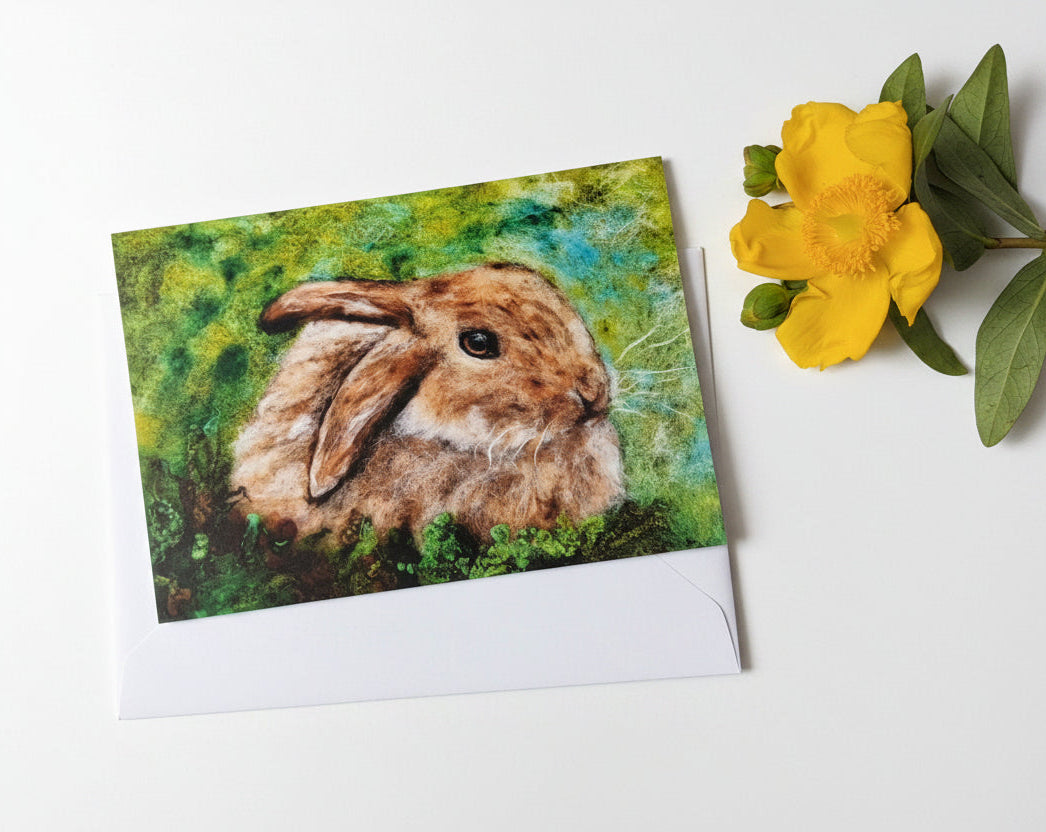 A square art print featuring a painted brown rabbit in a green and blue textured background, displayed on a white wooden surface next to a yellow flower.