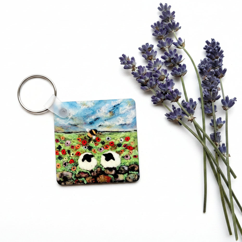 A square keychain with a colorful landscape illustration of sheep in a field under a blue sky, displayed on a textured gray surface next to purple lavender sprigs.