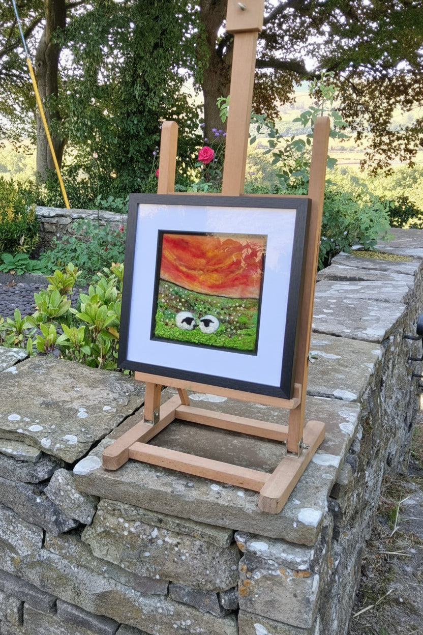 A framed art print featuring a colorful abstract design is displayed on a wooden easel outdoors next to a stone wall and trees.
