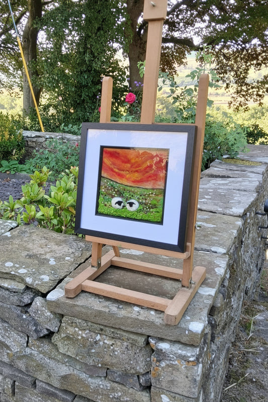 A framed art print featuring a colorful abstract design is displayed on a wooden easel outdoors next to a stone wall and trees.