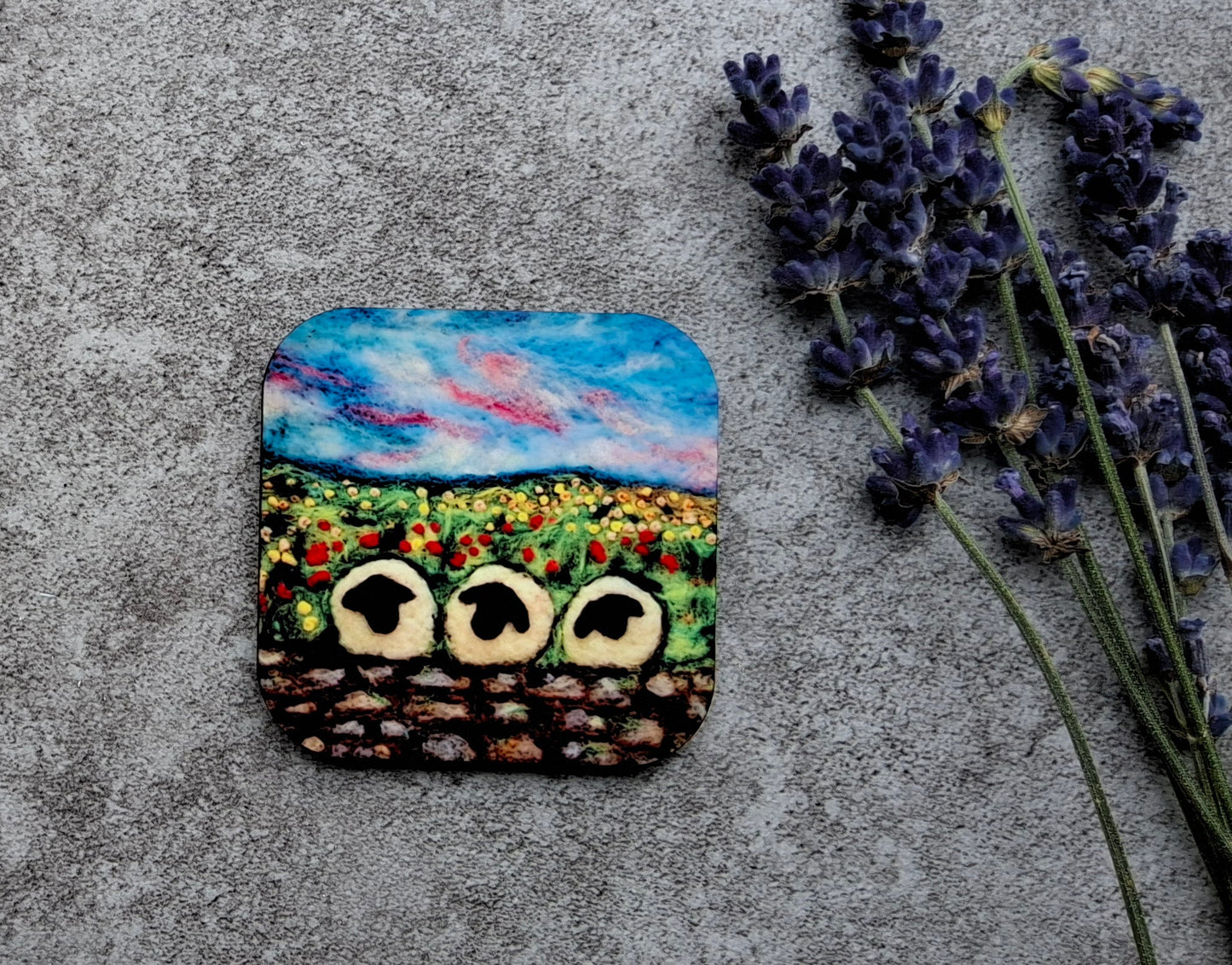 A square coaster featuring a colorful painted landscape of a sunflower field with three owl faces, styled on a textured gray concrete surface next to dried lavender sprigs.