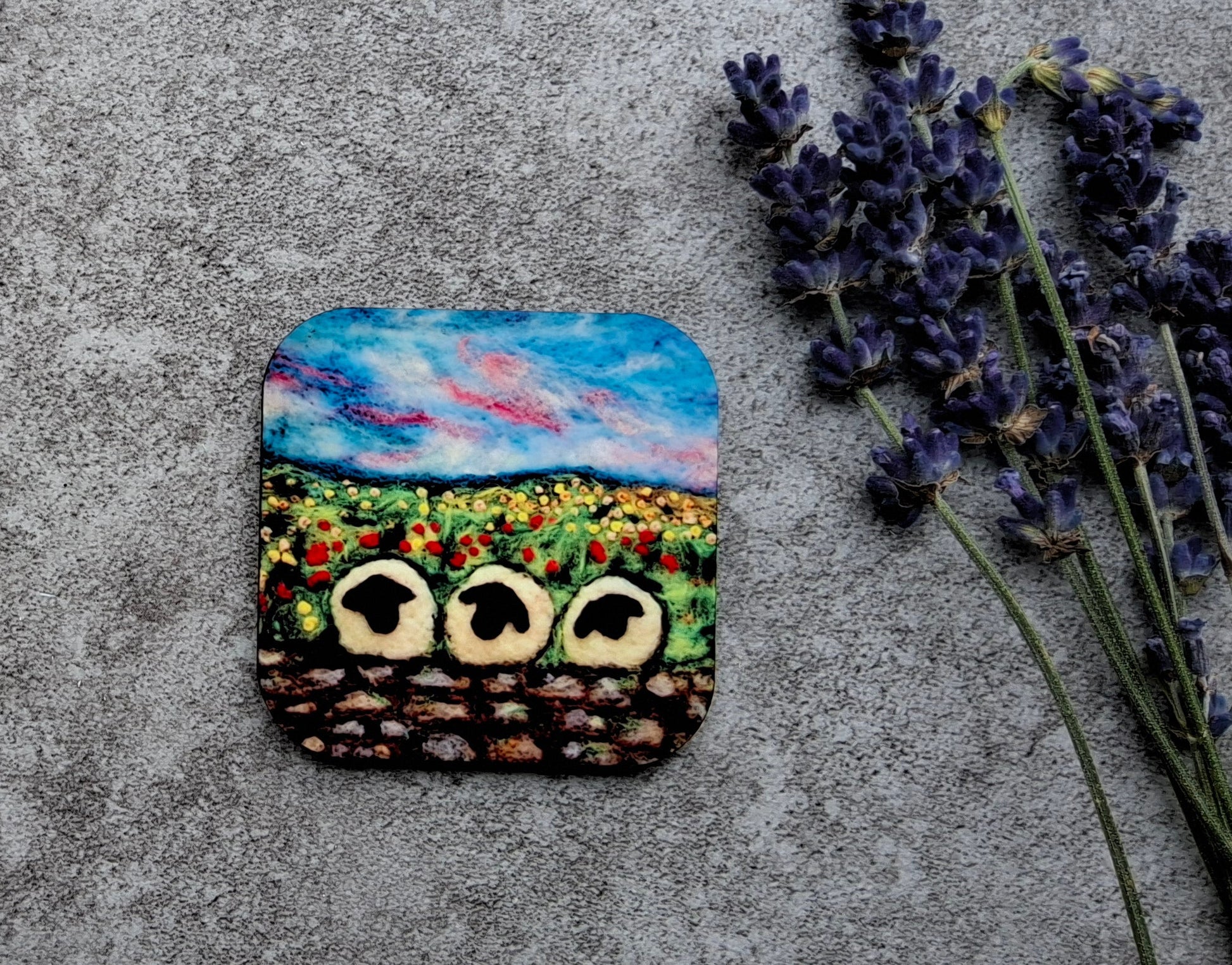 A square coaster featuring a colorful painted landscape of a sunflower field with three owl faces, styled on a textured gray concrete surface next to dried lavender sprigs.