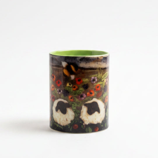 A small cylindrical cup or tumbler with a bright green interior and a dark exterior featuring a stylized face design, placed on a gray surface in front of a textured gray backdrop. A small green leafy sprig lies to the right.