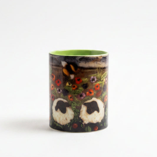 A small cylindrical cup or tumbler with a bright green interior and a dark exterior featuring a stylized face design, placed on a gray surface in front of a textured gray backdrop. A small green leafy sprig lies to the right.