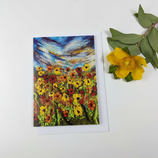 A square art print or card featuring an impressionistic painting of a flower field under a swirling blue sky, styled on a white wood surface next to a yellow flower and green leaves.
