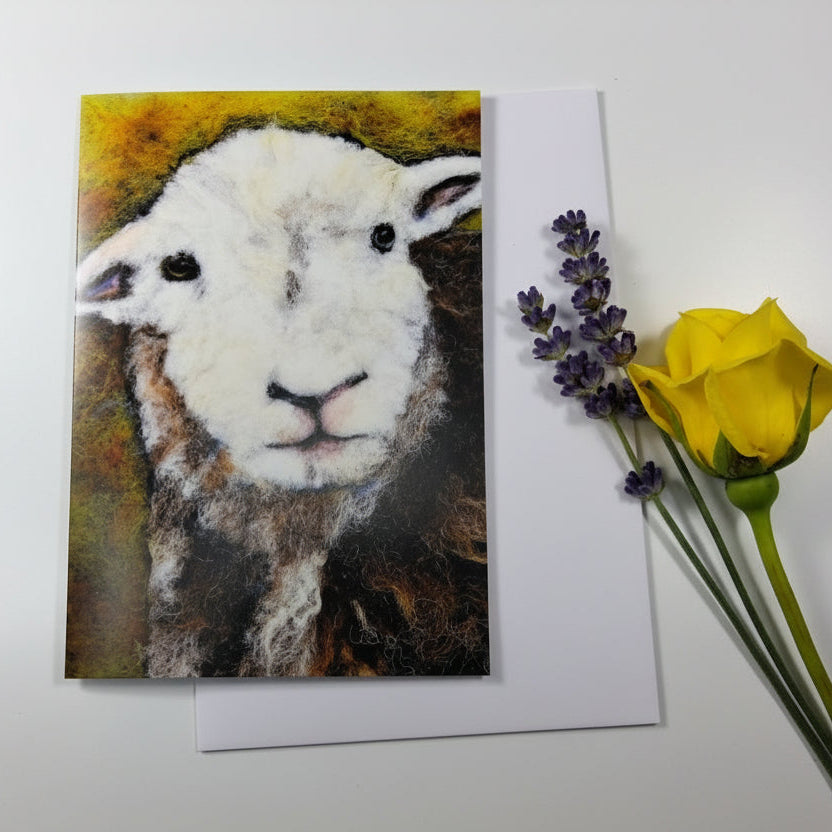A square art print or card featuring a painted white lamb face, placed on a white envelope over a whitewashed wood surface, styled with a yellow rose and lavender sprigs.