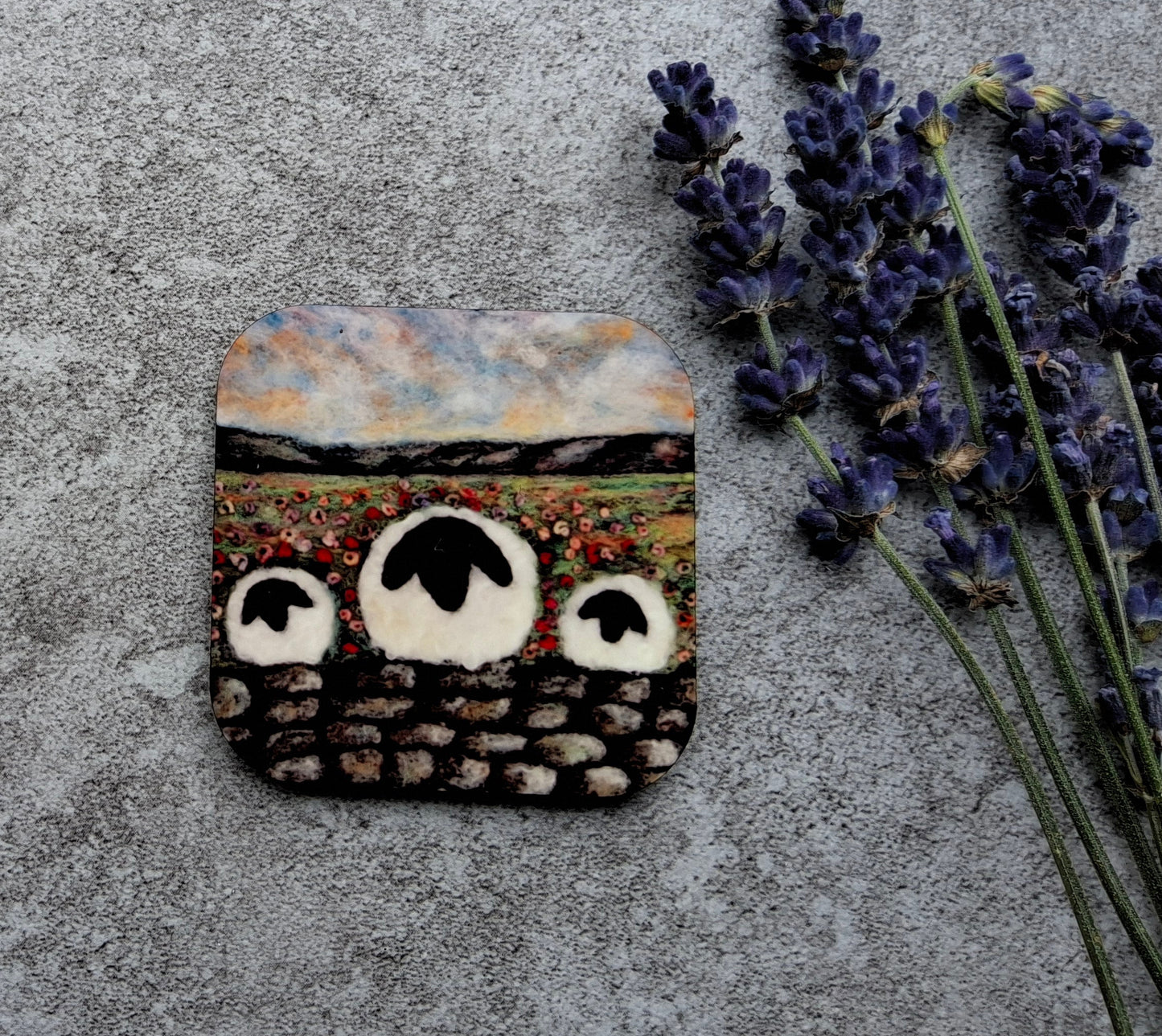 A square handmade stone mosaic piece depicting three sheep in a field, styled on a textured gray concrete surface next to sprigs of lavender.