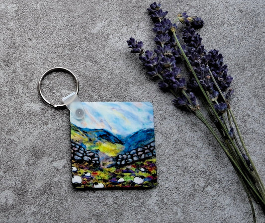 A small square keychain featuring a colorful landscape painting of mountains and lavender fields, displayed on a gray concrete surface next to dried lavender sprigs.