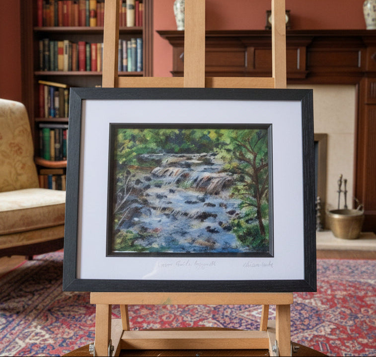 A framed landscape painting of a forest stream displayed on a wooden easel in a cluttered artist's studio workspace.