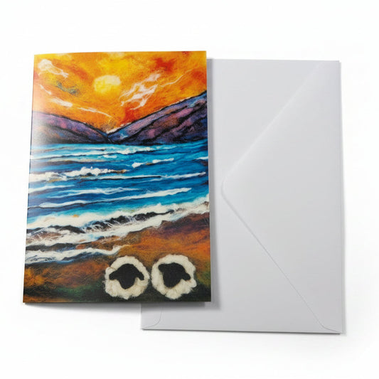 A square greeting card featuring a colorful landscape painting of a sunset over a blue ocean with white surf and two white ringed seals at the bottom, displayed on a white wooden surface next to an orange rose and a white envelope.