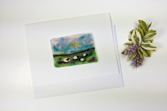 A white greeting card featuring a small, textured, hand-felted landscape painting with green fields, sheep, and a blue sky, displayed on a rustic white wooden table next to dried flowers.
