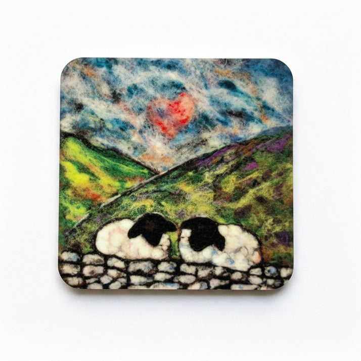 A square coaster featuring a painted pastoral landscape with two black-and-white sheep in the foreground, green hills, and a cloudy sky, photographed on a gray textured surface next to a red rose.