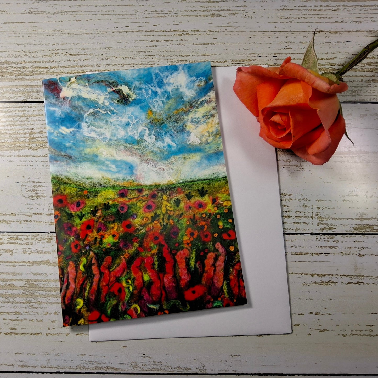 A square art print or greeting card featuring an impressionistic landscape painting of a flower field under a blue sky, placed on a white envelope over a distressed white wood surface with an orange rose beside it.