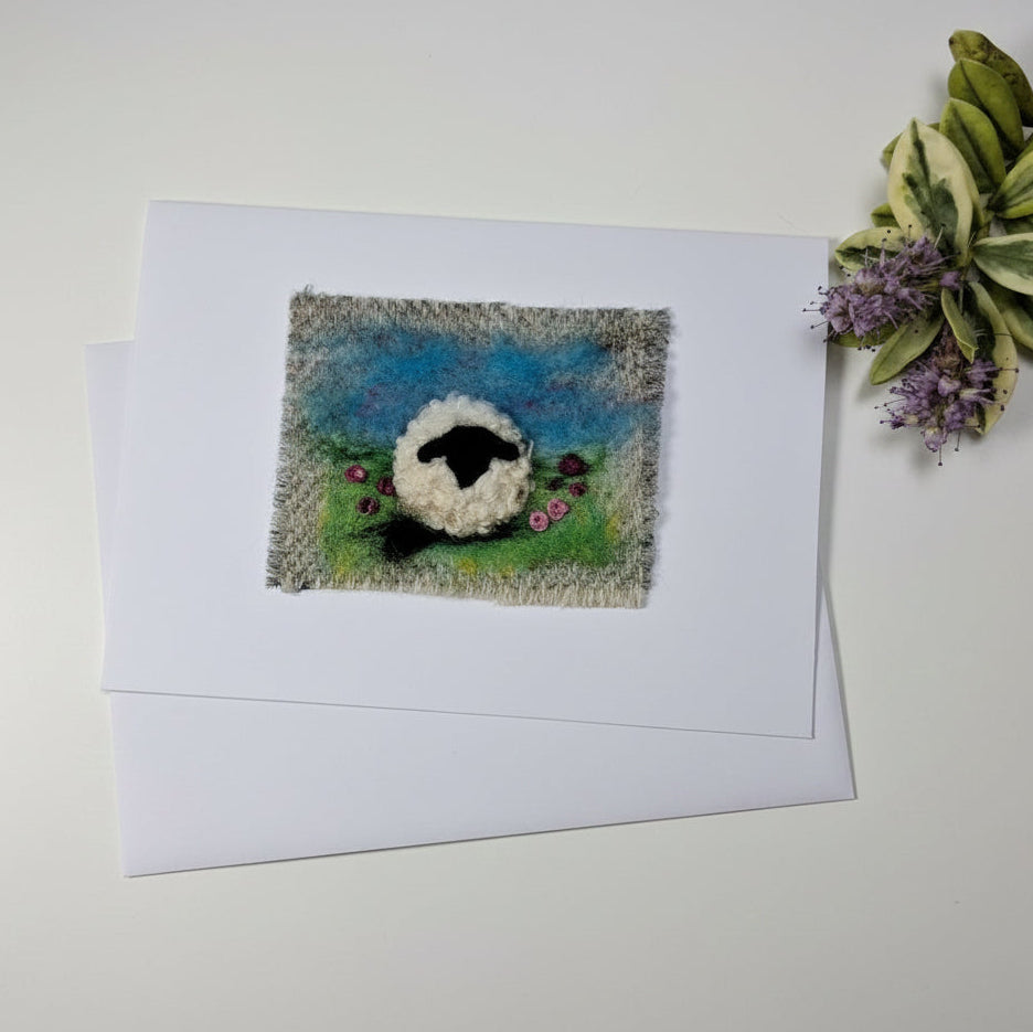 A square handmade felted wool art piece depicting a white sheep with a black face on a blue and green textured background, displayed on a white card over a distressed white wood tabletop with decorative greenery.