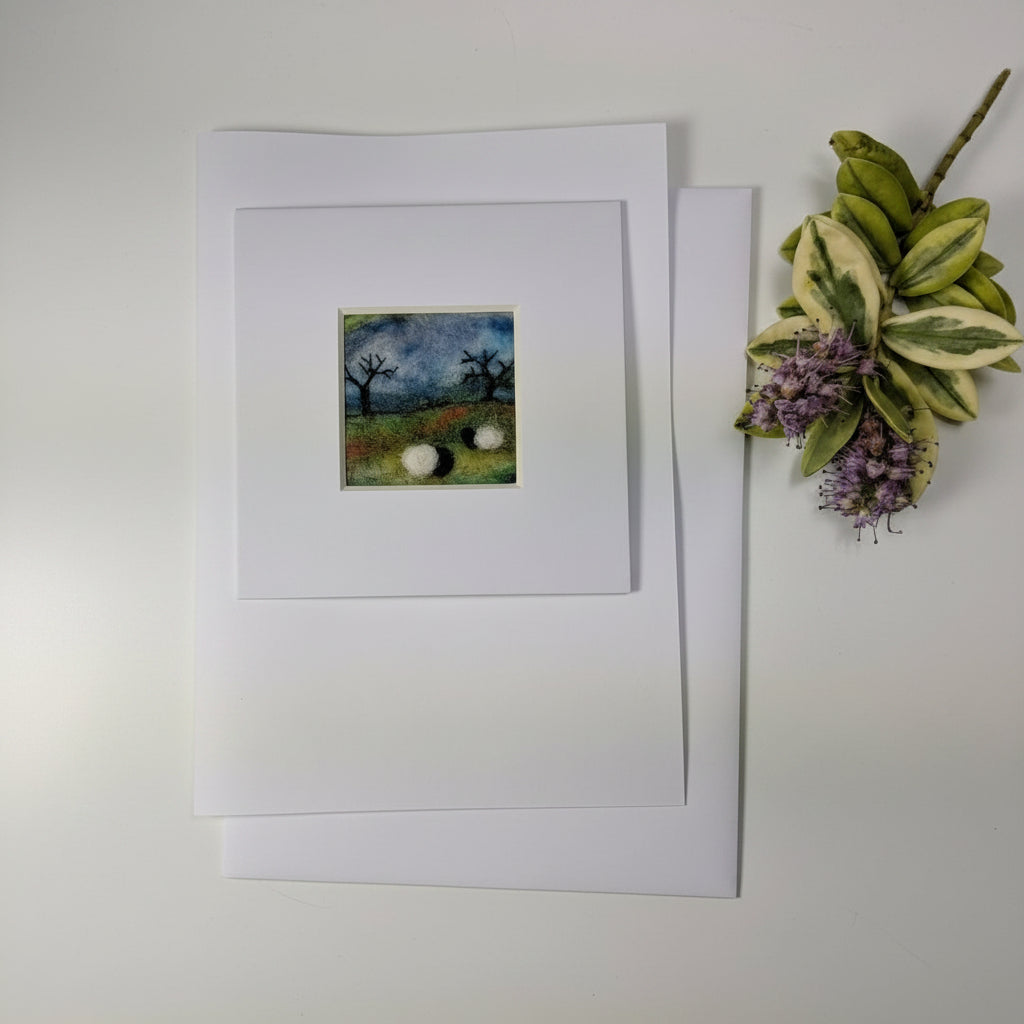 A square white art print or greeting card with a wide white mat, featuring a small central landscape illustration of trees and sheep, displayed on a rustic whitewashed wooden surface next to a decorative plant sprig.
