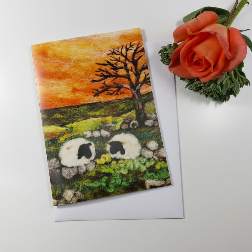 A rectangular art print or card showing a painted pastoral landscape with two white sheep, a bare tree, rocks, and an orange-yellow sky, 