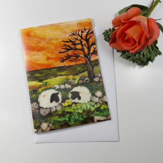 A rectangular art print or card showing a painted pastoral landscape with two white sheep, a bare tree, rocks, and an orange-yellow sky, 