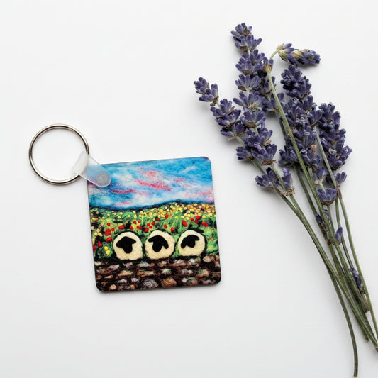 A square acrylic keychain featuring a colorful painted landscape of sunflowers and a blue sky, shown on a textured gray concrete surface next to dried lavender sprigs.