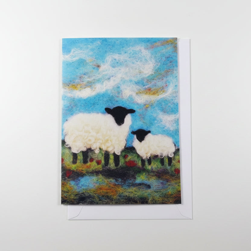 A square art print or card featuring a painted scene of two white sheep with black heads standing in a grassy field under a blue sky, placed on a white surface next to a pink flower.