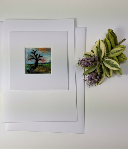 A square art print featuring a dark tree silhouette against a colorful sky, matted on white paper and displayed on a rustic white wooden table next to a decorative plant sprig.