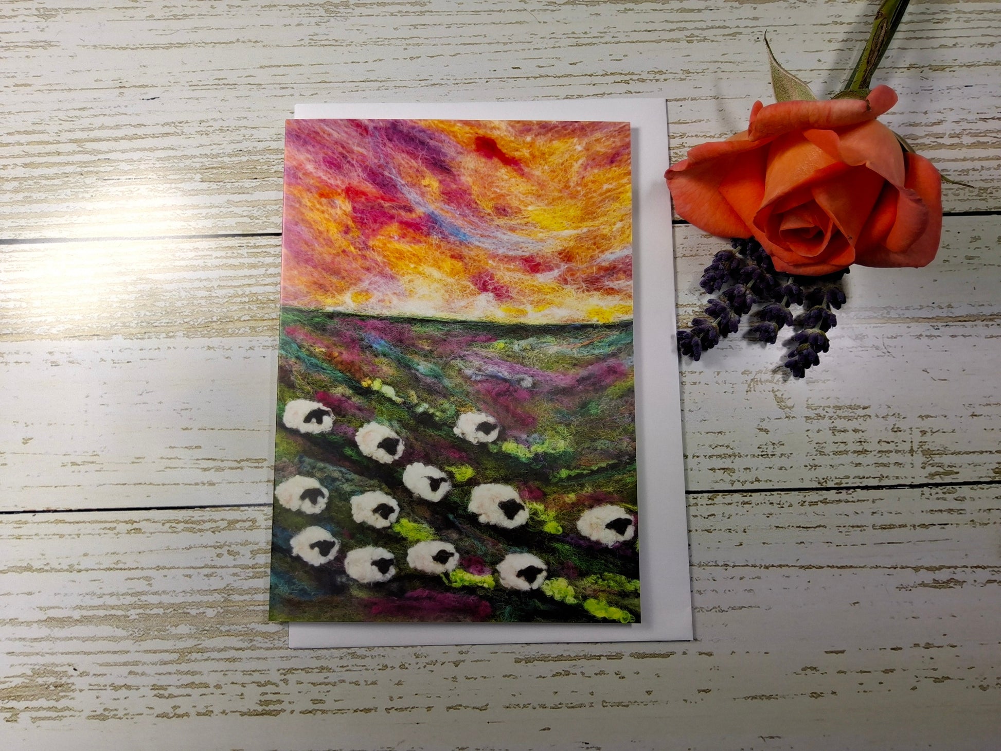 A vertical art print or card featuring a colorful landscape painting of sheep in a field under a vibrant sunset sky, displayed on a white wood background next to an orange rose and purple grapes.