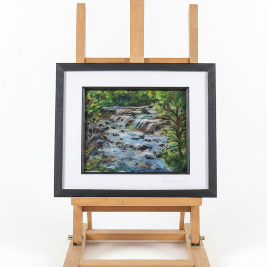 A framed art print of a forest stream displayed on a small wooden easel in a cluttered craft room or studio workspace.