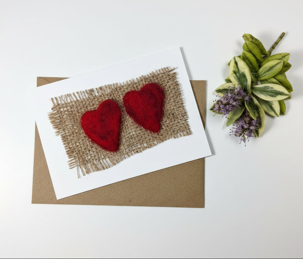 A handmade greeting card featuring two red felt hearts on a piece of burlap, placed on top of a brown kraft envelope on a distressed white wood background.