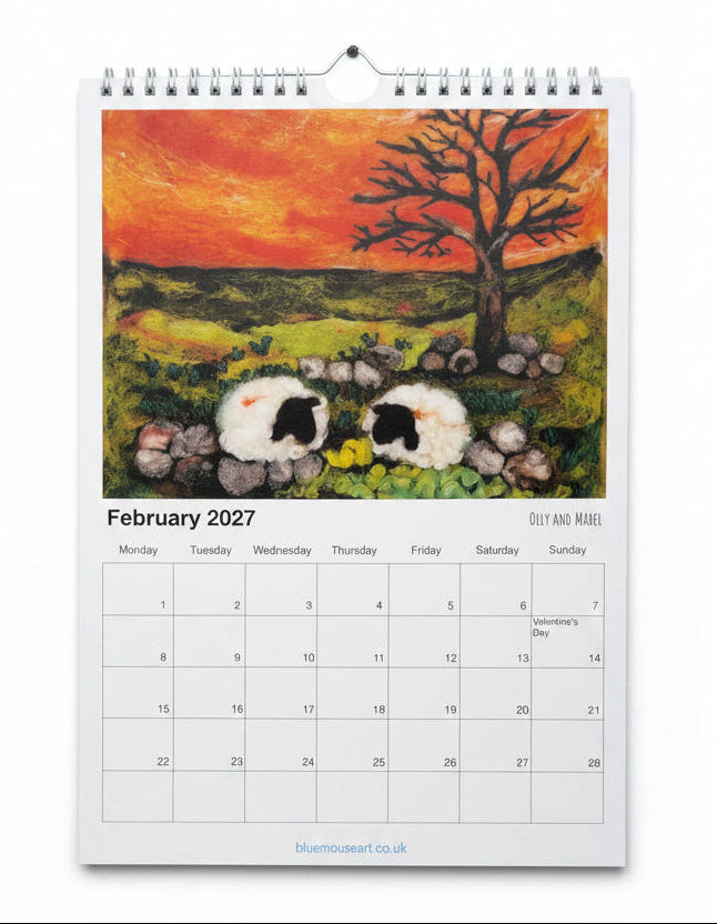 A spiral-bound February 2027 desk calendar page featuring a colorful hand-painted style illustration of two sheep in a field under a tree with an orange sky, photographed against a whitewashed wood background.