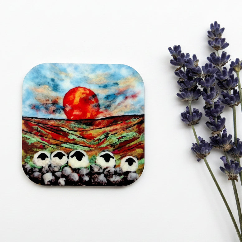 A square coaster with rounded corners featuring a colorful painted landscape of sheep on a hillside under a red sun, styled on a gray textured surface next to purple lavender stems.