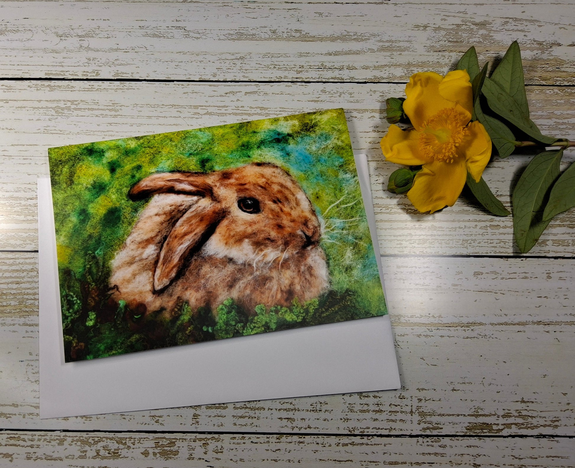 A square art print or greeting card featuring a painted brown rabbit in green grass, placed on a white envelope over a whitewashed wood surface, with a yellow flower beside it.