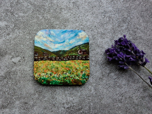 A square coaster featuring a colorful landscape painting of a field under a blue sky, placed on a textured gray surface next to a sprig of purple lavender.