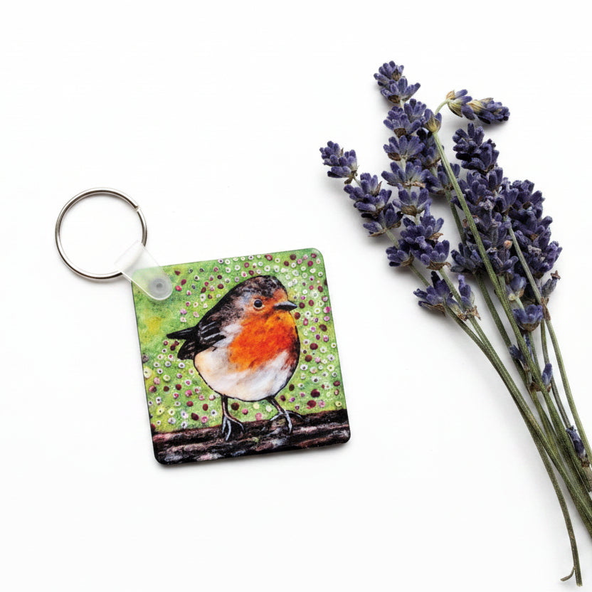 A square keychain featuring a painted robin bird on a green textured background, shown on a gray concrete surface next to dried lavender sprigs.