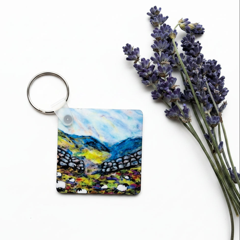 A square acrylic keychain featuring a colorful landscape painting of a mountain valley, styled on a textured gray concrete surface next to sprigs of purple lavender.